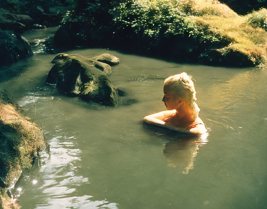 Person relaxing in serene forest pond with rocks and trees.