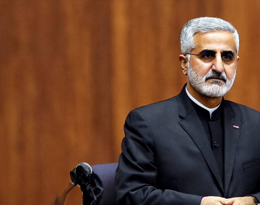 Salt-and-pepper haired man in black suit without tie at microphone against wooden backdrop