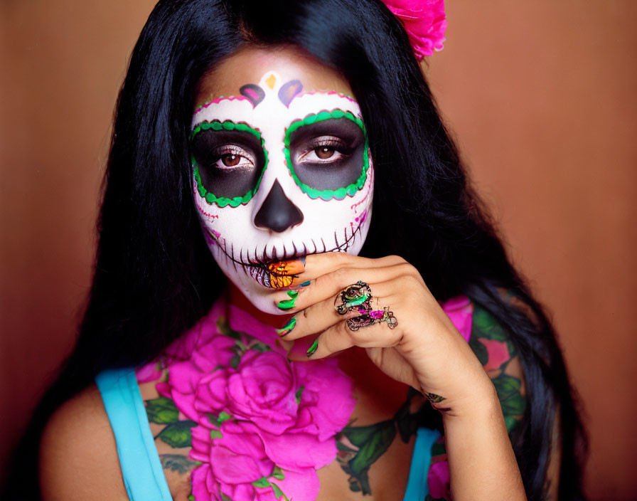 Colorful Day of the Dead makeup on woman with pink flower and tattoos against brown background