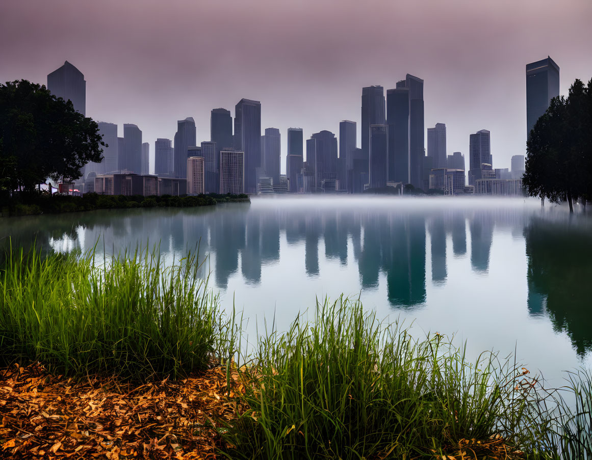 Tranquil cityscape reflected in calm lake with green grass, under hazy sky