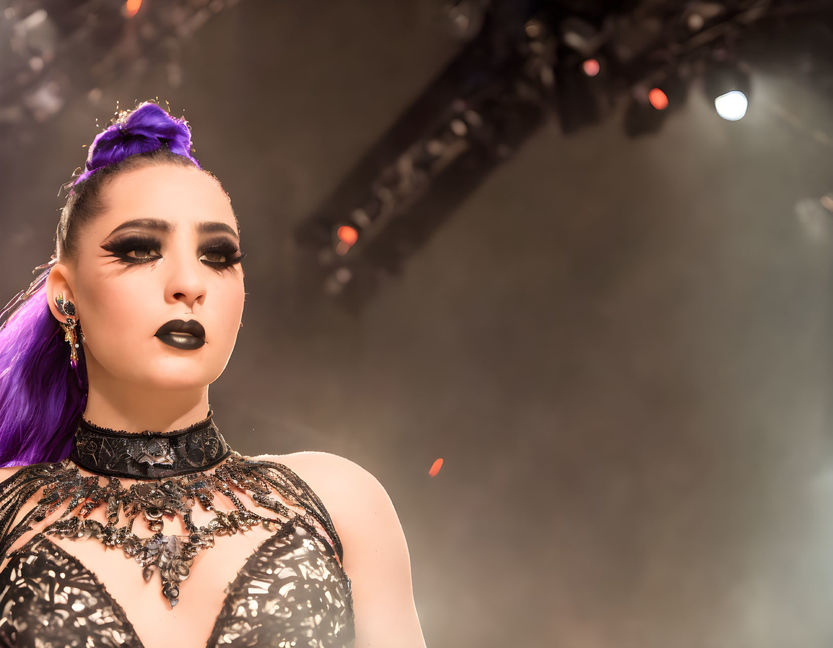 Purple-haired woman in topknot, dark makeup, and black lace outfit performs on stage with background