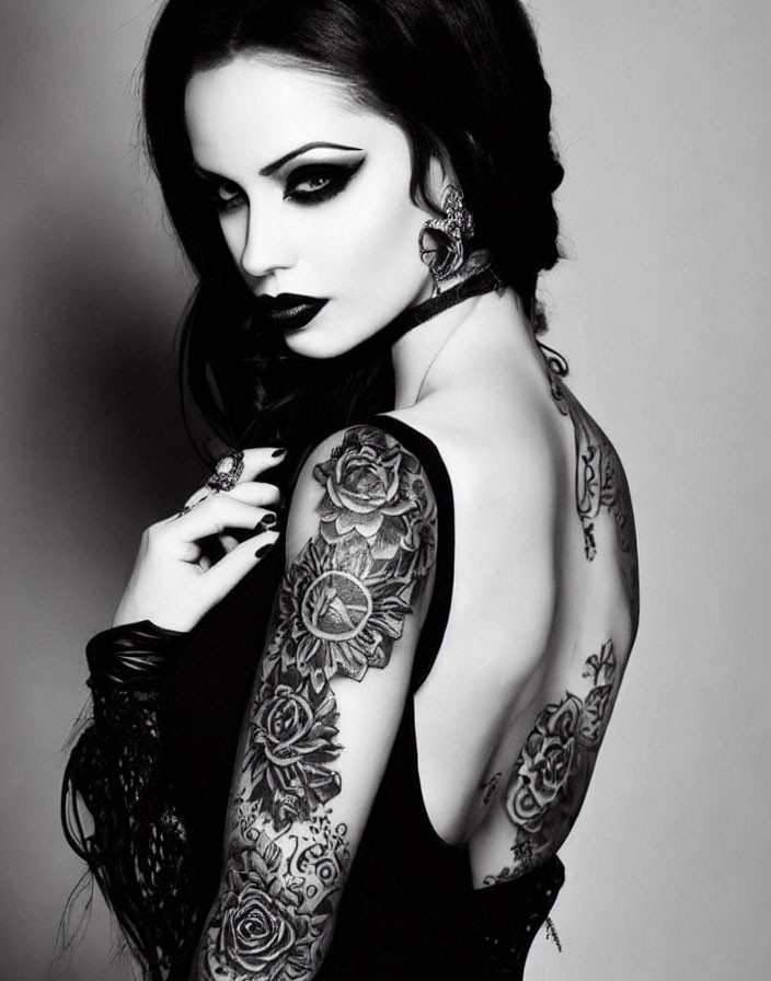 Monochrome portrait of woman with dark makeup, floral tattoos, sleeveless top, and dramatic earrings