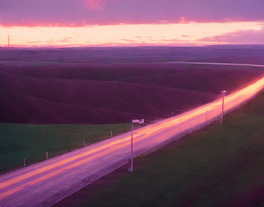 Vibrant sunset colors over rolling hills and car light trails on highway.