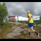 Runner in Yellow Top Jogging on Rocky Terrain with Red Cabin and Stormy Sky