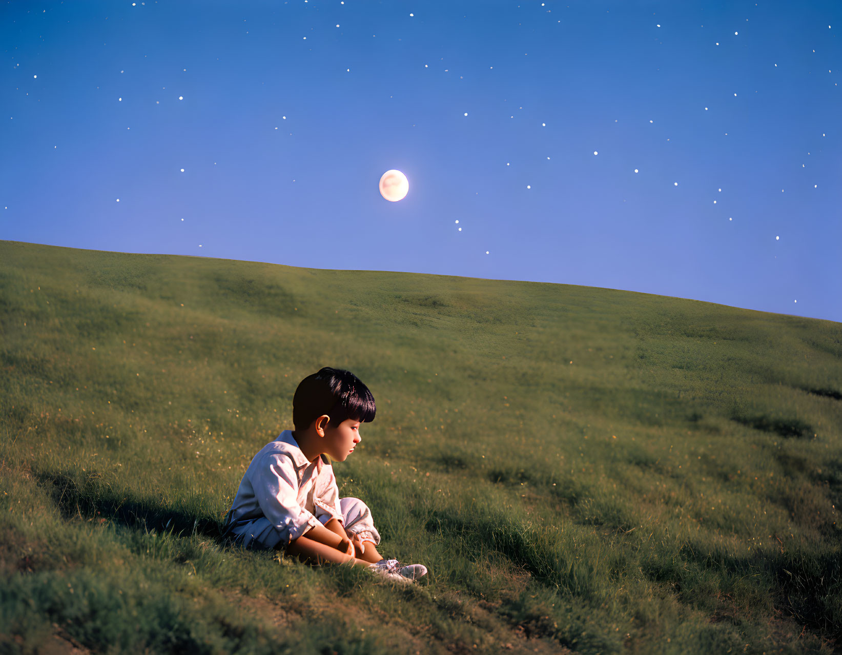 Young boy sitting on grassy hill under twilight sky with stars and moon.