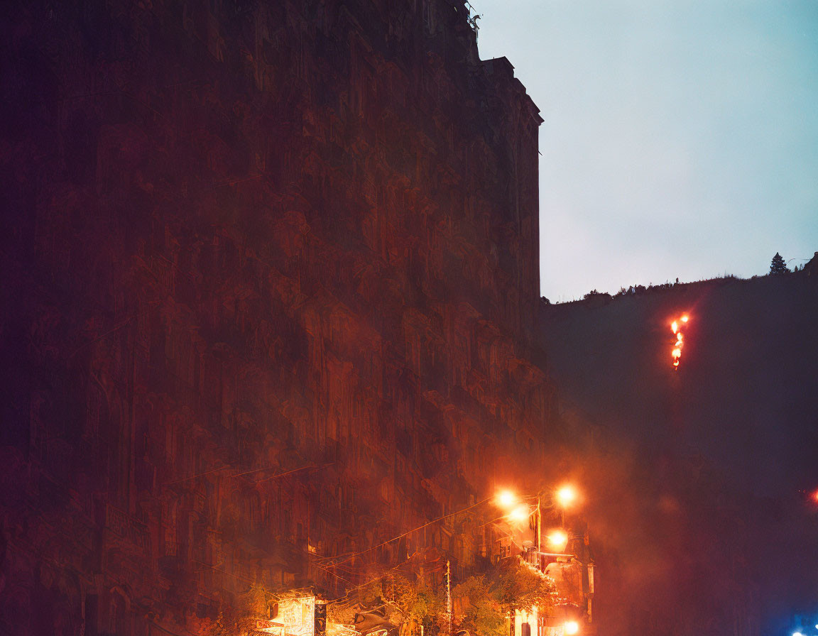 Night scene: illuminated streetlights, towering rocky cliff, faint cross.