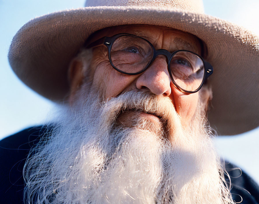 Elderly man with white beard, glasses, hat, and coat against blue sky