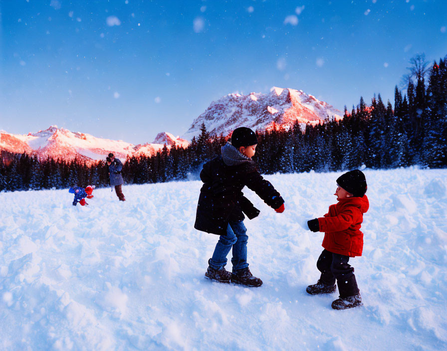 Children playing in snow with mountain backdrop, adult and child in distance, snowflakes in air