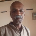 Elderly man in beige shirt with thin mustache and glasses by thatched-roof hut