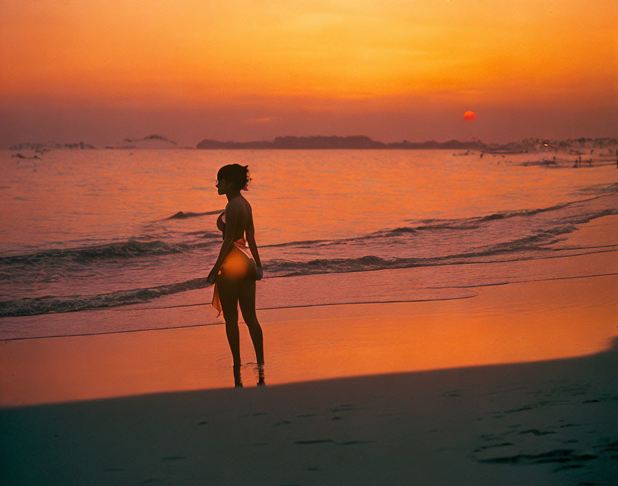 Person's silhouette on beach at sunset with orange glow.