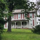 White two-story house with porch in overgrown greenery under cloudy sky