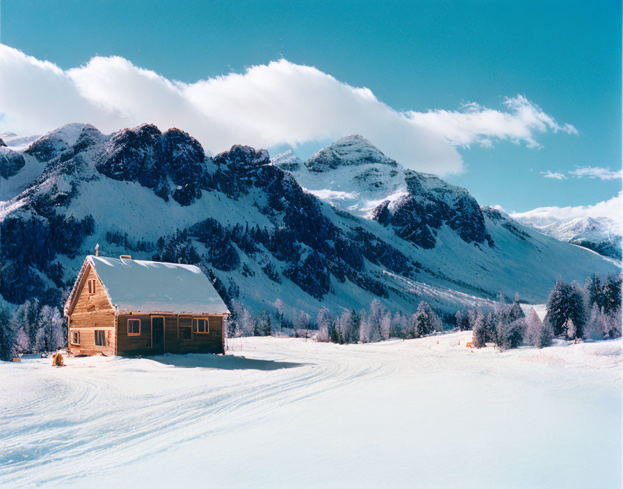 Snow-covered wooden cabin amidst snowy trees and mountains under blue sky.