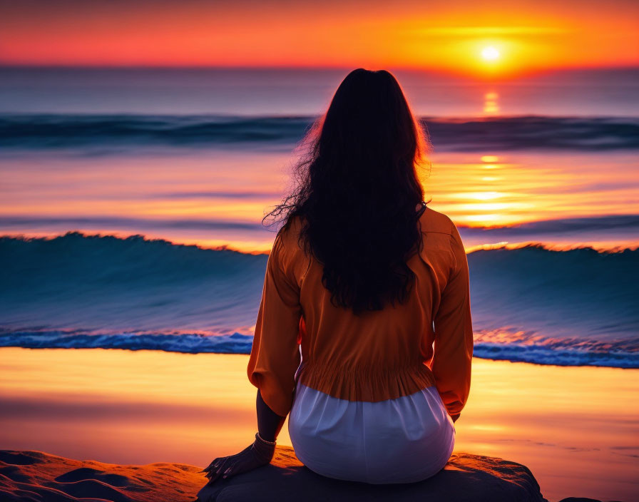 Long-haired person sitting on beach at sunset gazes at sun-ocean horizon