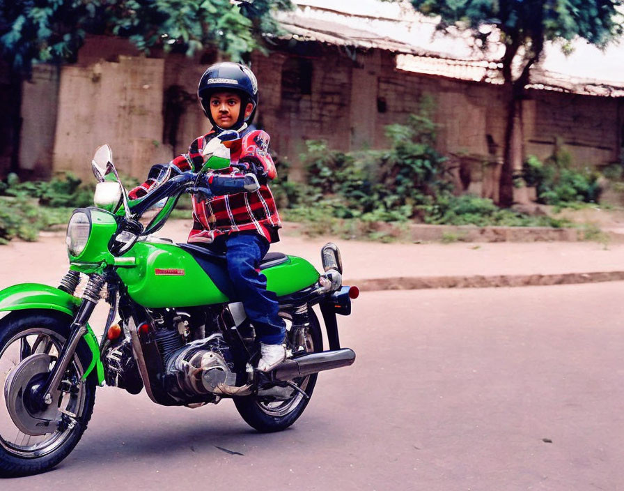 Child in helmet on green motorcycle by road buildings & trees