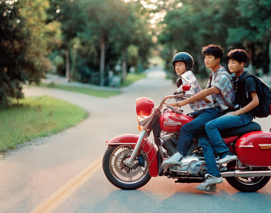 Three people on red motorcycle with helmets and backpacks on tree-lined road at golden hour