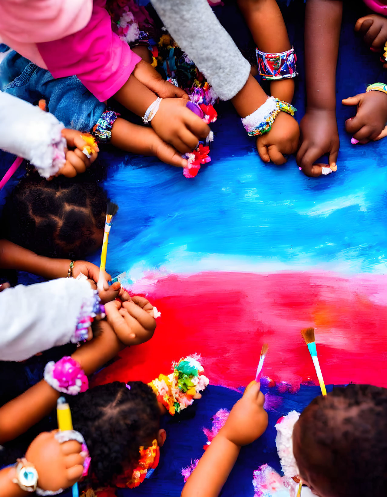 Colorful bracelets on children's hands painting with bright blue and red paints on canvas in group activity