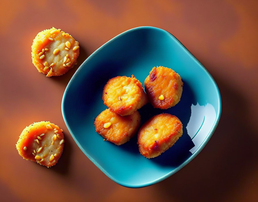 Teal bowl with golden-brown fried snacks and sesame seeds on brown surface