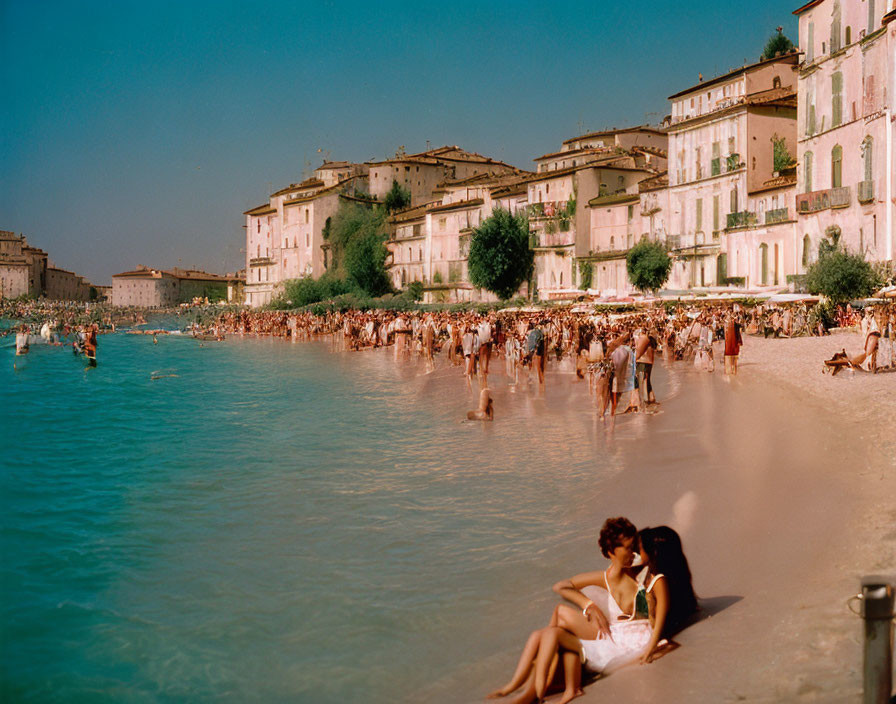 Crowded beach scene with people and pastel buildings under clear sky