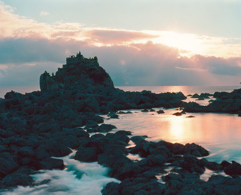 Tranquil sunset scene with rocky outcrop and small structure by calm sea