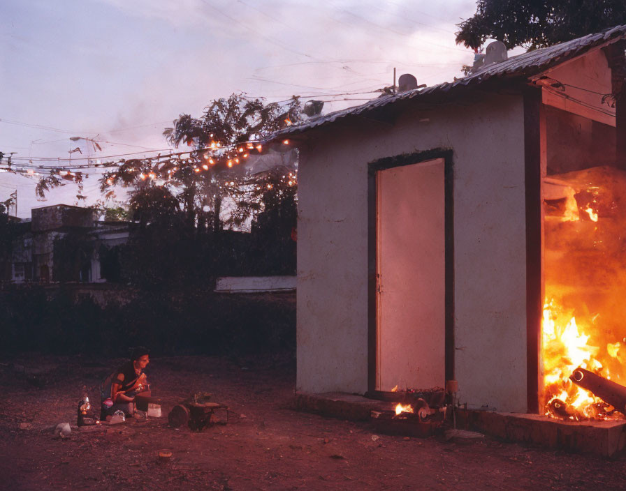 Person cooking beside small building with room on fire under dusky sky