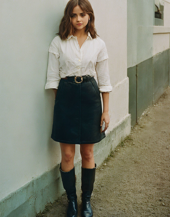 Person in white shirt, black skirt, and boots against wall