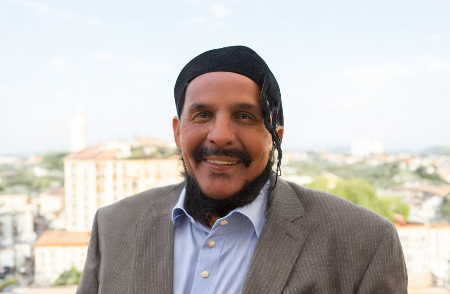 Smiling man in black head covering and blue attire with cityscape background