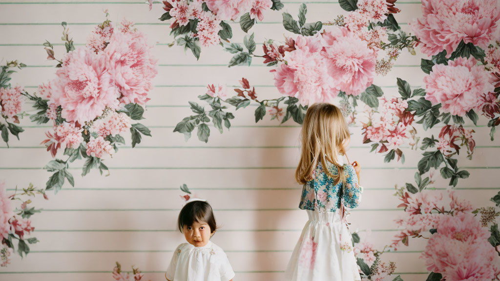 Children in front of floral wallpaper, one standing, the other shorter, facing away.