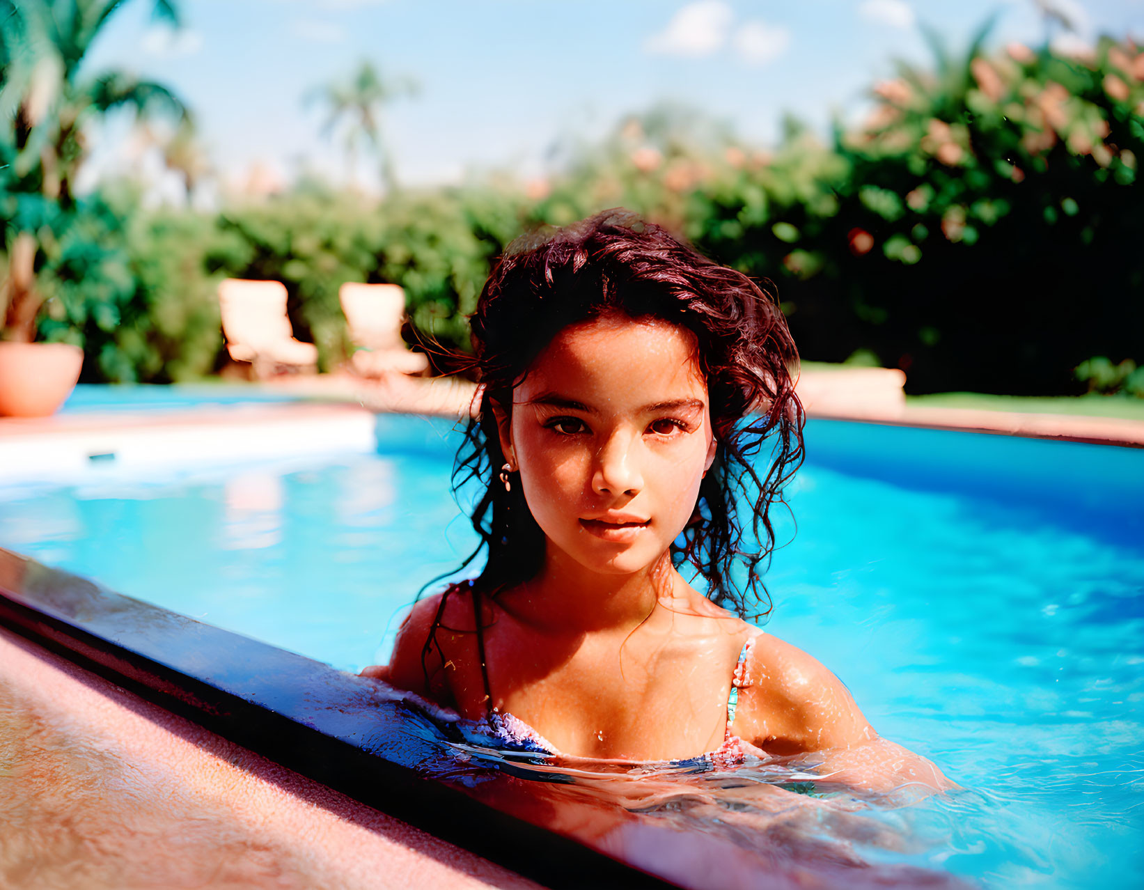 Young girl at swimming pool with wet hair, greenery backdrop & blue sky