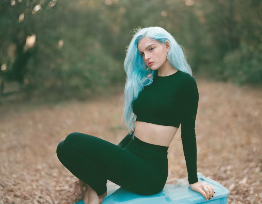 Blue-haired woman on stool in forest with fallen leaves wearing black clothes