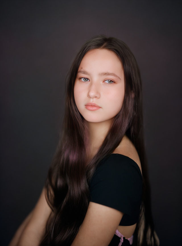 Young person with long brown hair and blue eyes in black top on dark background