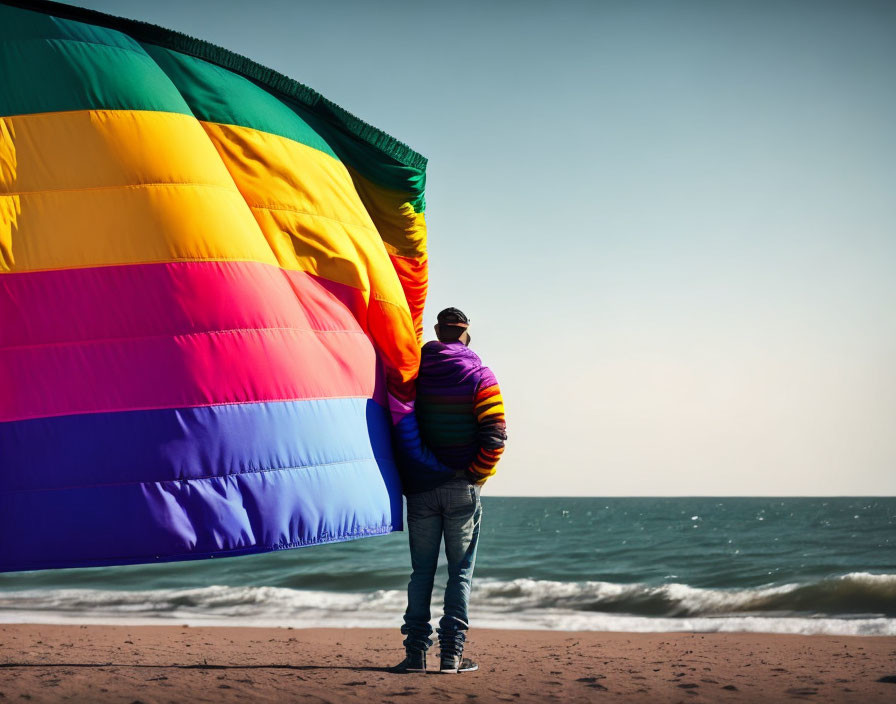 Person in multi-colored jacket on beach with rainbow kite.