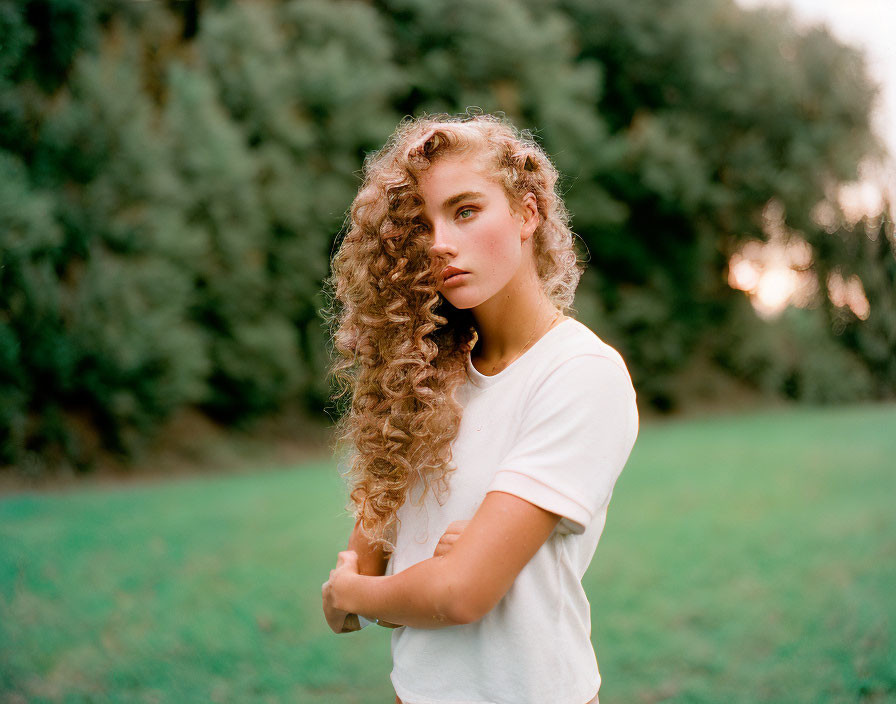 Long Curly-Haired Person in White T-Shirt Standing in Field with Trees