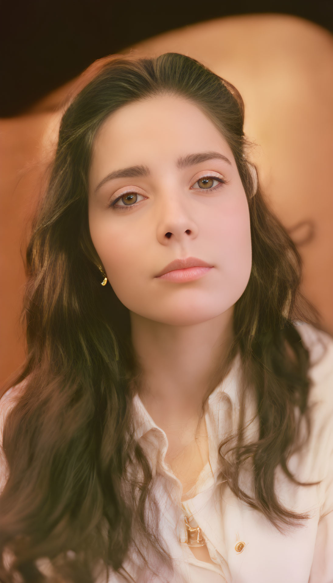 Woman with dark hair and green eyes in white blouse and earrings on warm backdrop