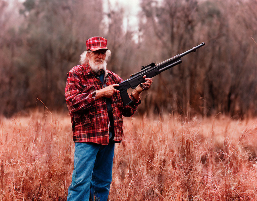 Elderly man with a beard holding a rifle in a field with dry grass and trees