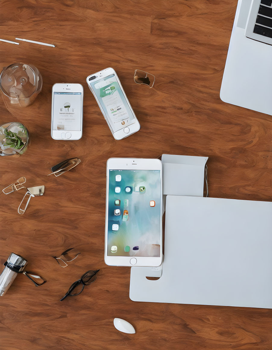 Modern workspace setup with laptop, tablet, smartphones, glasses, pen, headphones, and drink on wooden