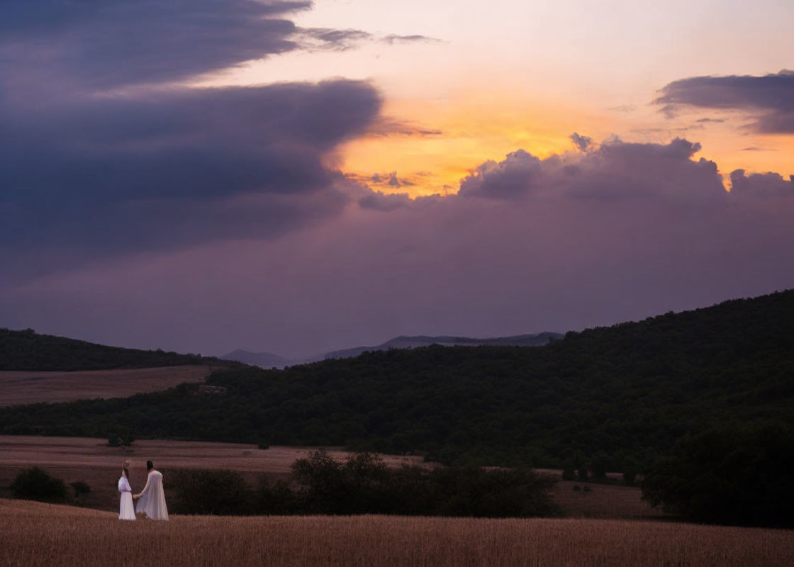 Two individuals in white attire amidst a sunset landscape with colorful sky and rolling hills.