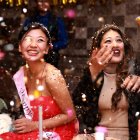 Group celebrating pink-themed birthday party with cake, hats, and balloons. Smiling people in festive setting