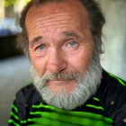 Elderly man portrait with white beard, green shirt, blue jacket, and leafy background