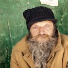 Elderly man with long beard in beret and coat by cracked green wall