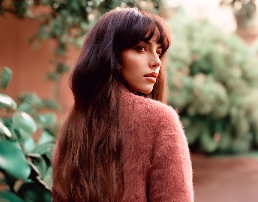 Dark-haired woman in pink fluffy jacket gazes at camera in garden setting