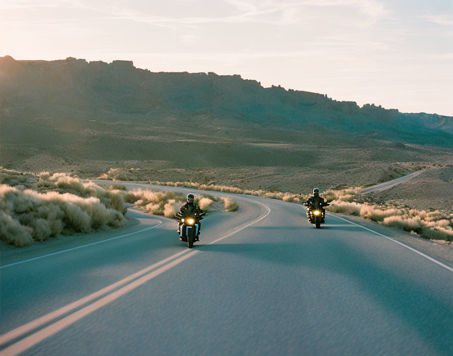 Two motorcyclists on curvy desert road at dawn with mountains in background