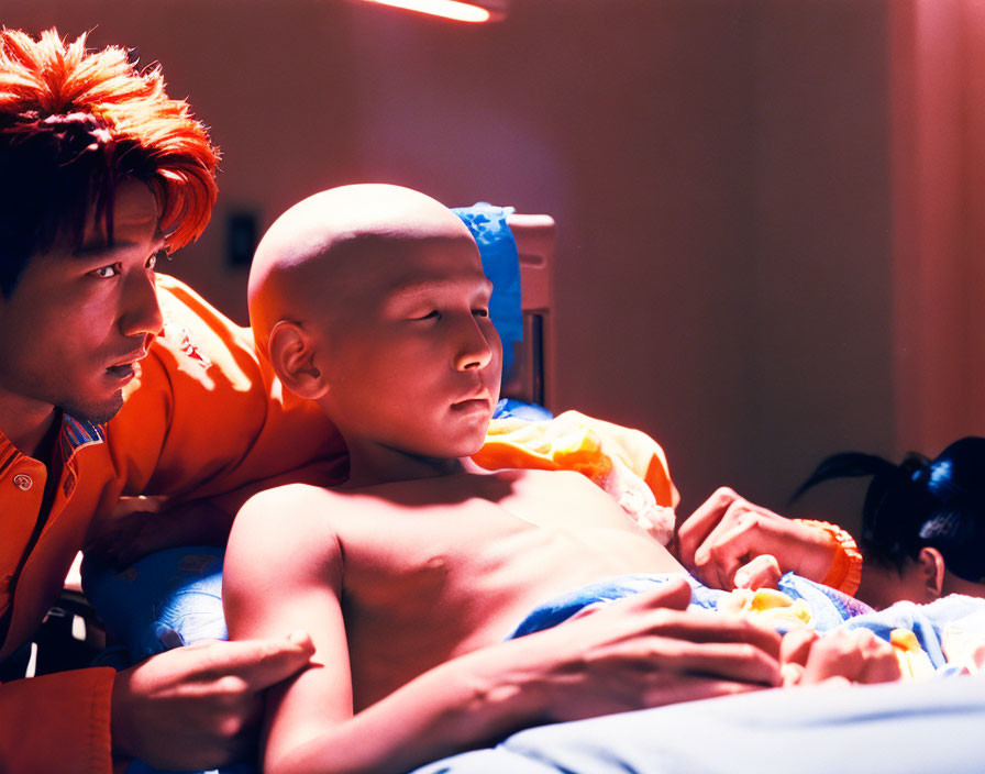 Man in orange jacket gazes at bald child in dimly lit room