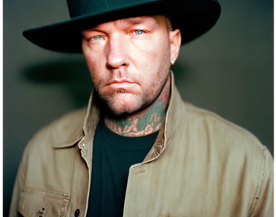 Tattooed man in black hat and khaki jacket with stern expression