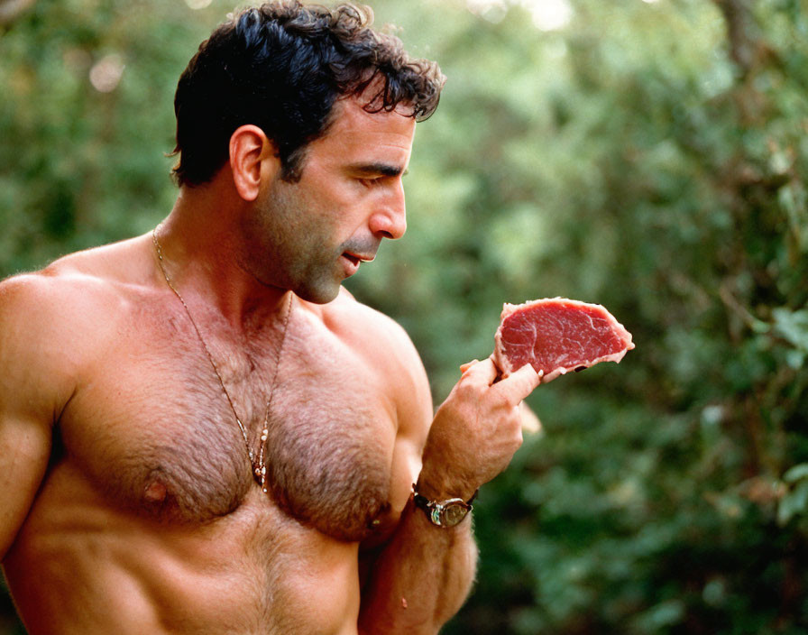 Muscular man with stubble holding raw steak in wooded area