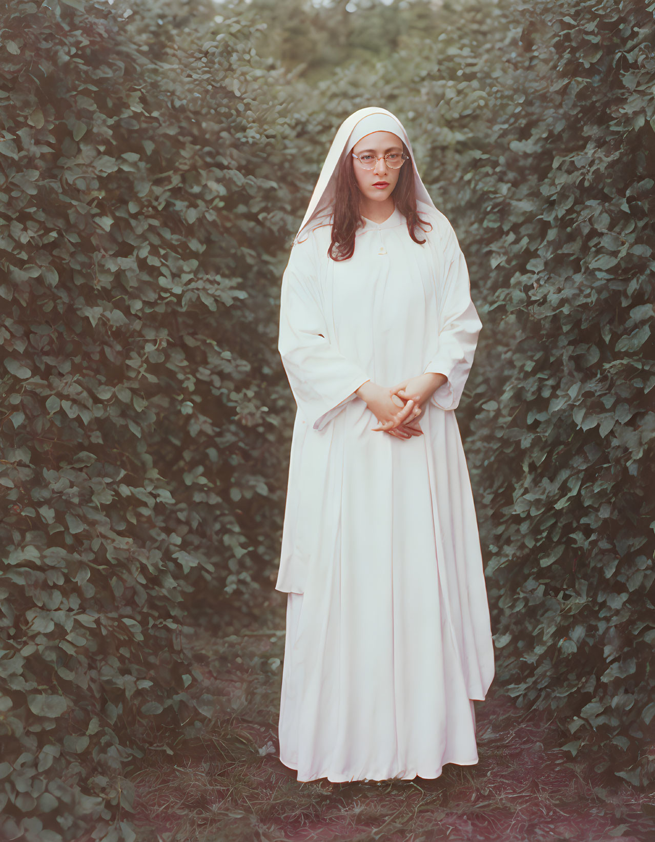 Woman in white habit and veil standing solemnly in green shrubbery