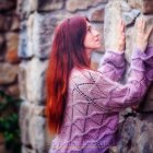 Red-haired woman in purple floral dress reading book against flower wall