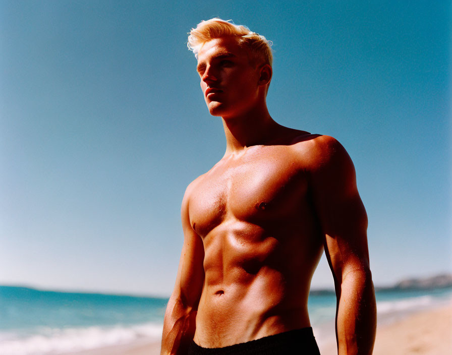 Muscular shirtless man with blond hair on beach against clear blue sky