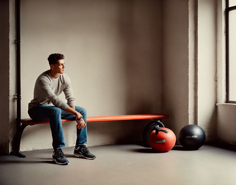 Person sitting on orange bench near medicine balls in room with large windows