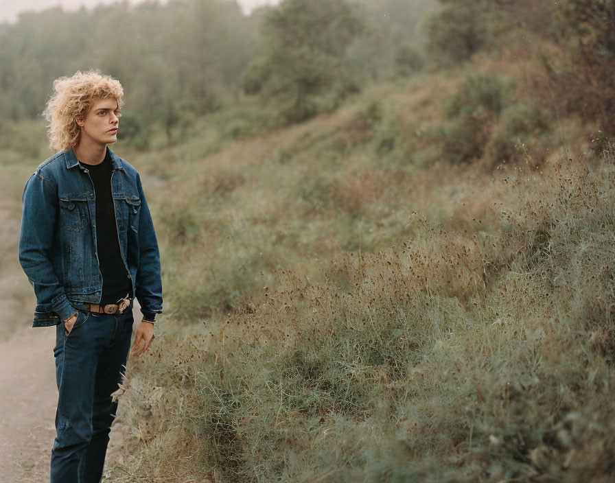 Young person with blonde curly hair in denim jacket gazes on grassy hillside