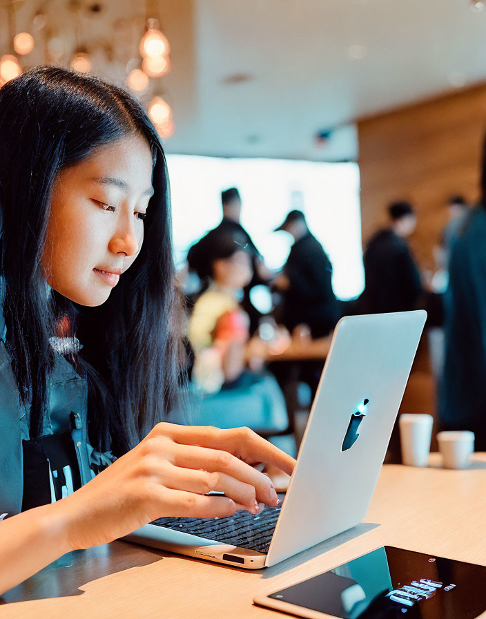 Woman working on laptop in busy café with people and smartphone.